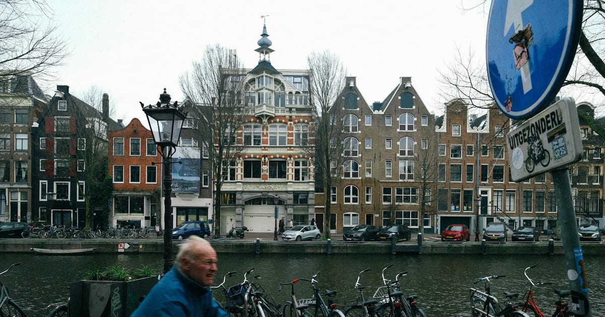 An older man cycling past a row of parked bicycles beside a canal and terraced houses.