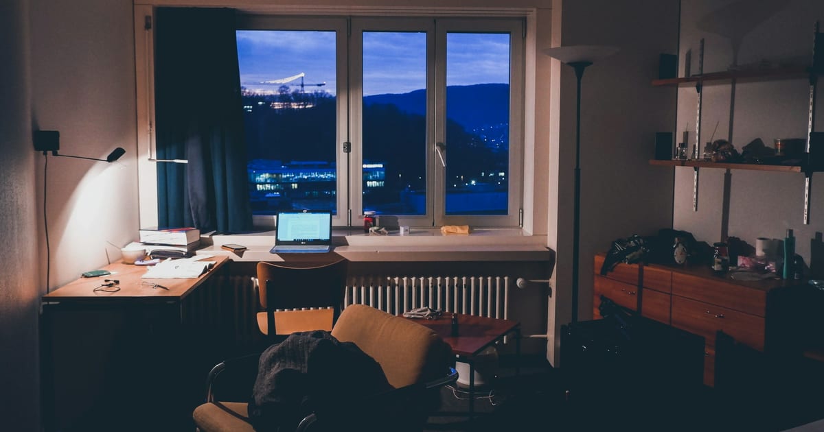 A laptop open on a desk in a dark home office, city lights visible through the window.