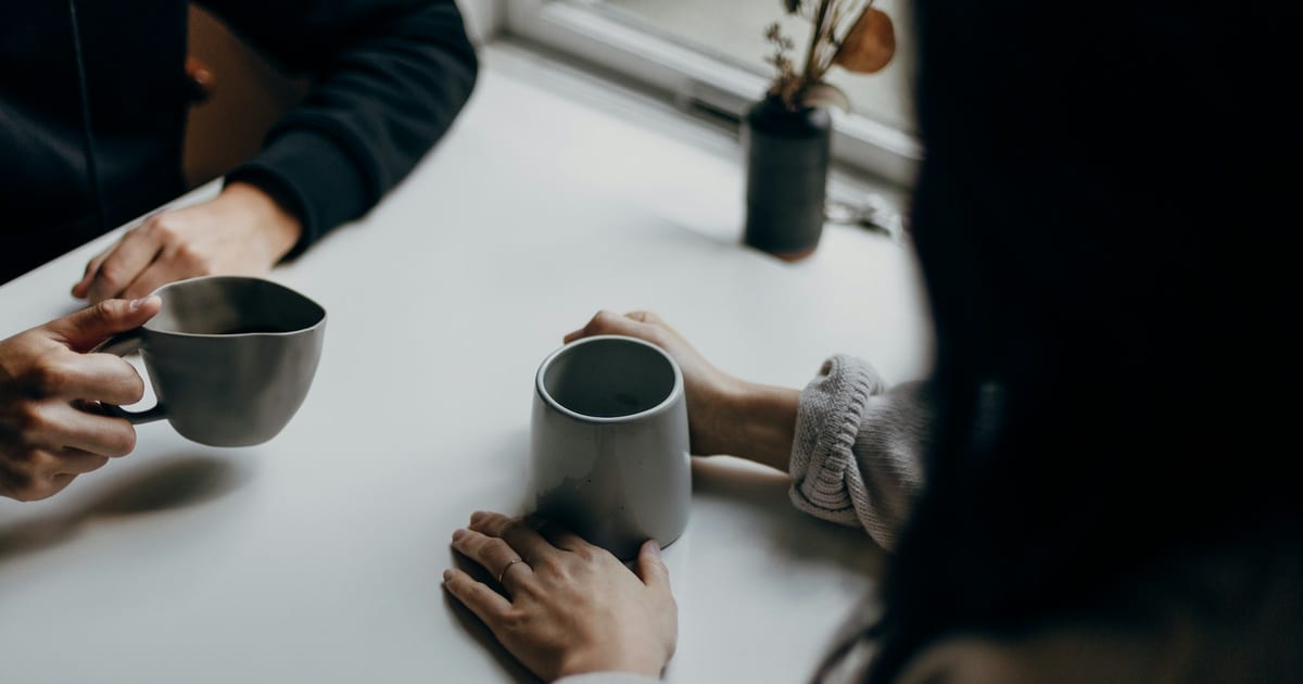 Two people sitting across a table, each holding a grey mug.