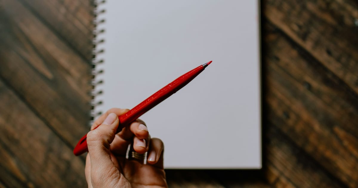 A hand holding a red pen above a blank spiral notebook on a wooden table.
