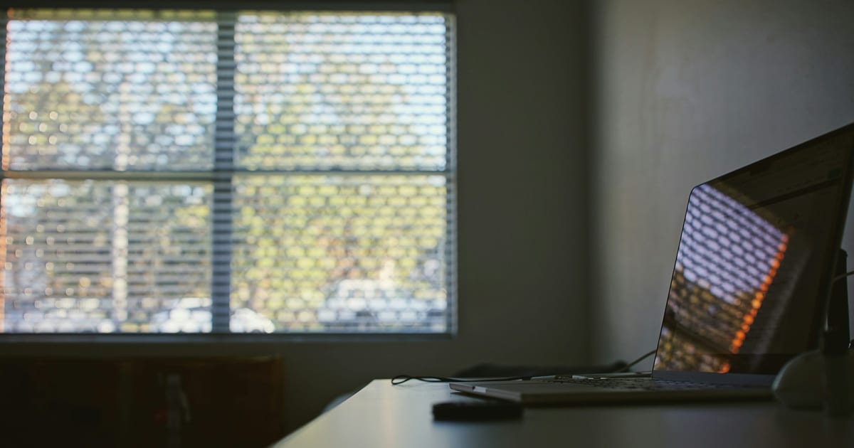 A laptop open on a desk in a dim room, in front of a window with closed blinds.